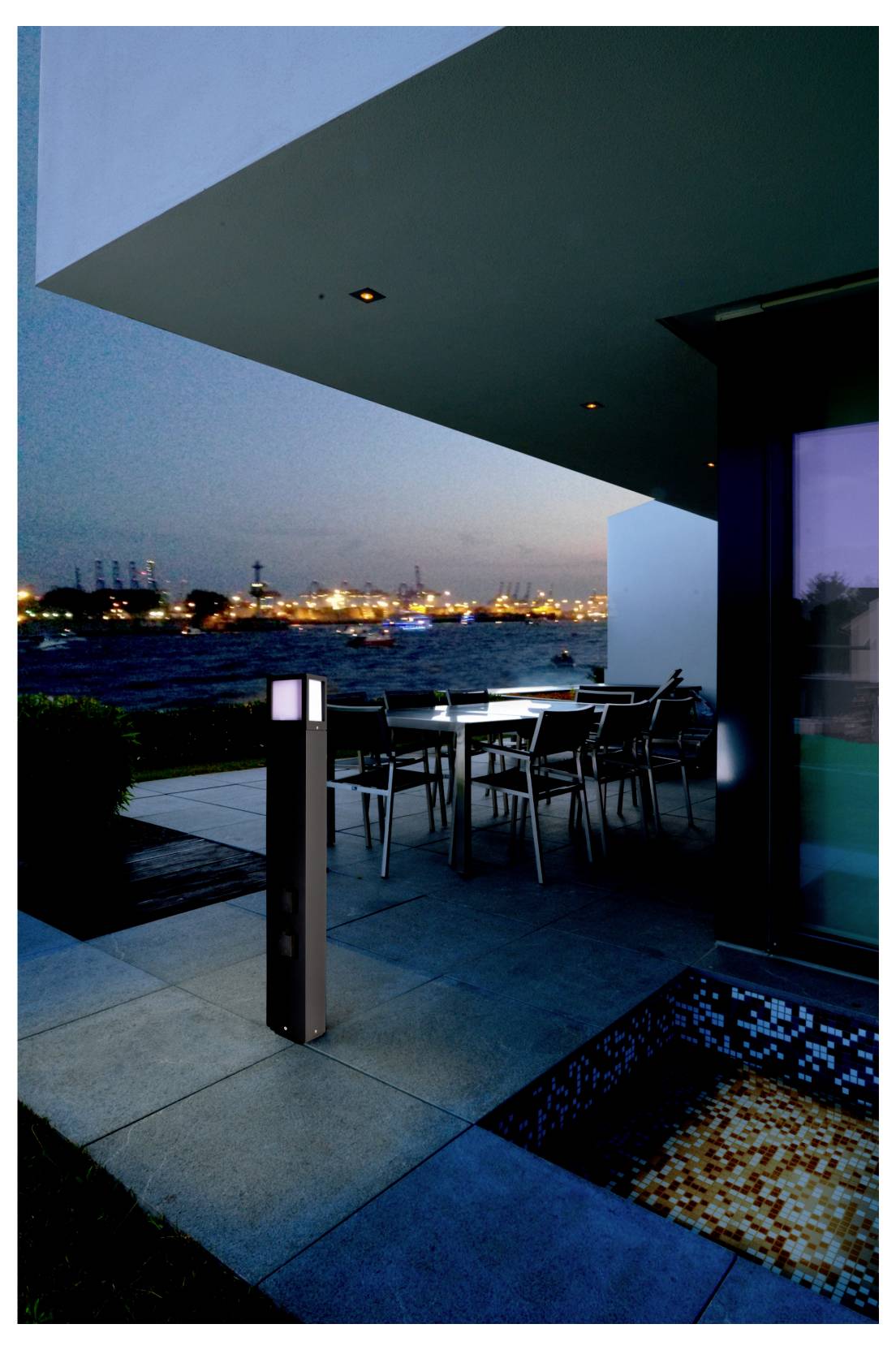 Moderne Terrasse am Wasser in der Dämmerung mit Esstischen im Freien, einer beleuchteten Laterne und den Lichtern der Hafenstadt im Hintergrund.