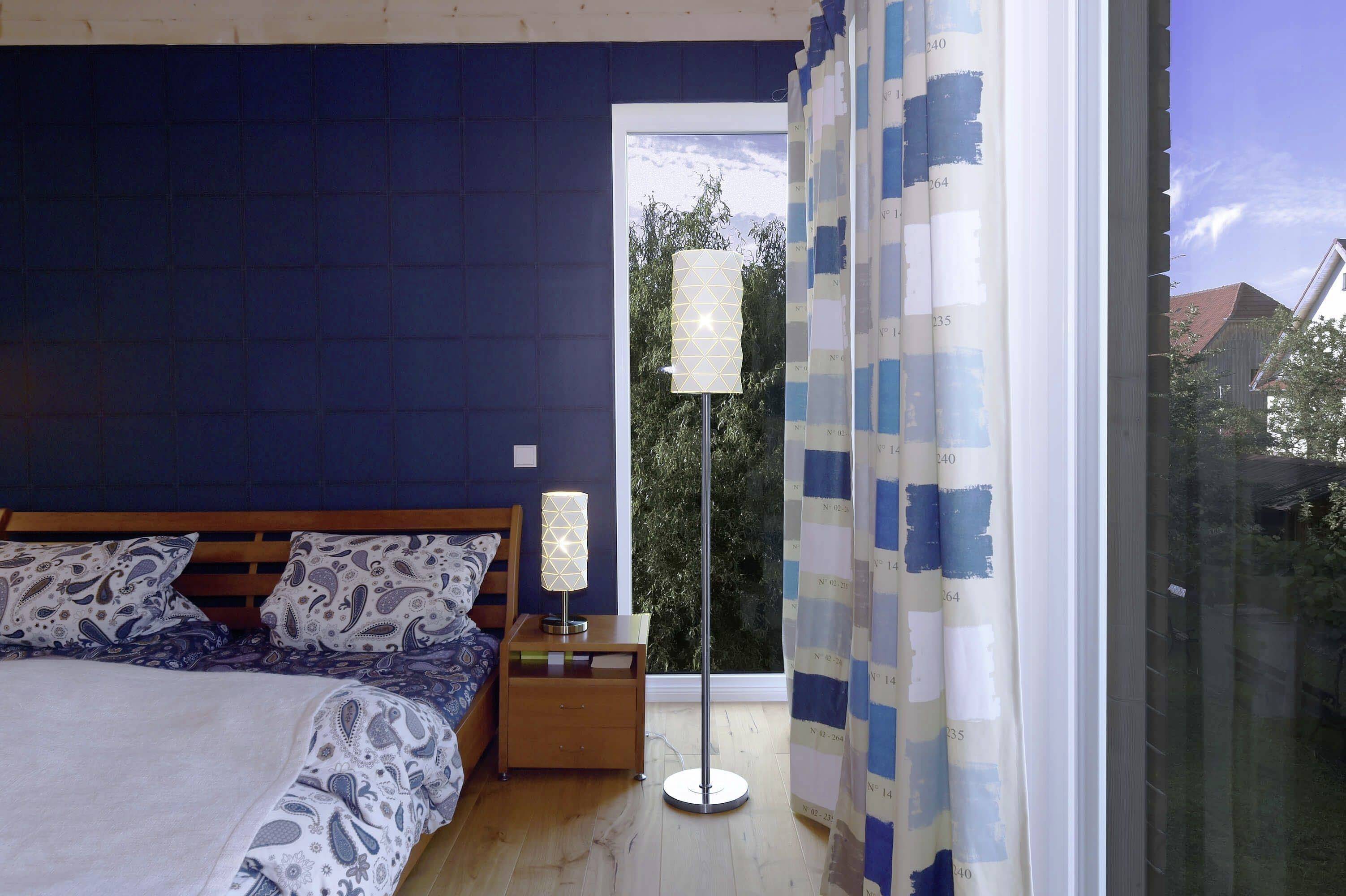Schlafzimmer mit Doppelbett, Nachttisch mit Lampe und einem großen Fenster mit Blick auf Bäume, blau-weiße Vorhänge zur Seite geschoben.