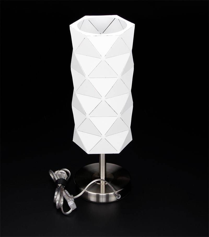 Weiße Tischlampe mit geometrischem, facettiertem Schirm auf einem silbernen Ständer vor schwarzem Hintergrund. Kabel ist sichtbar am Boden.