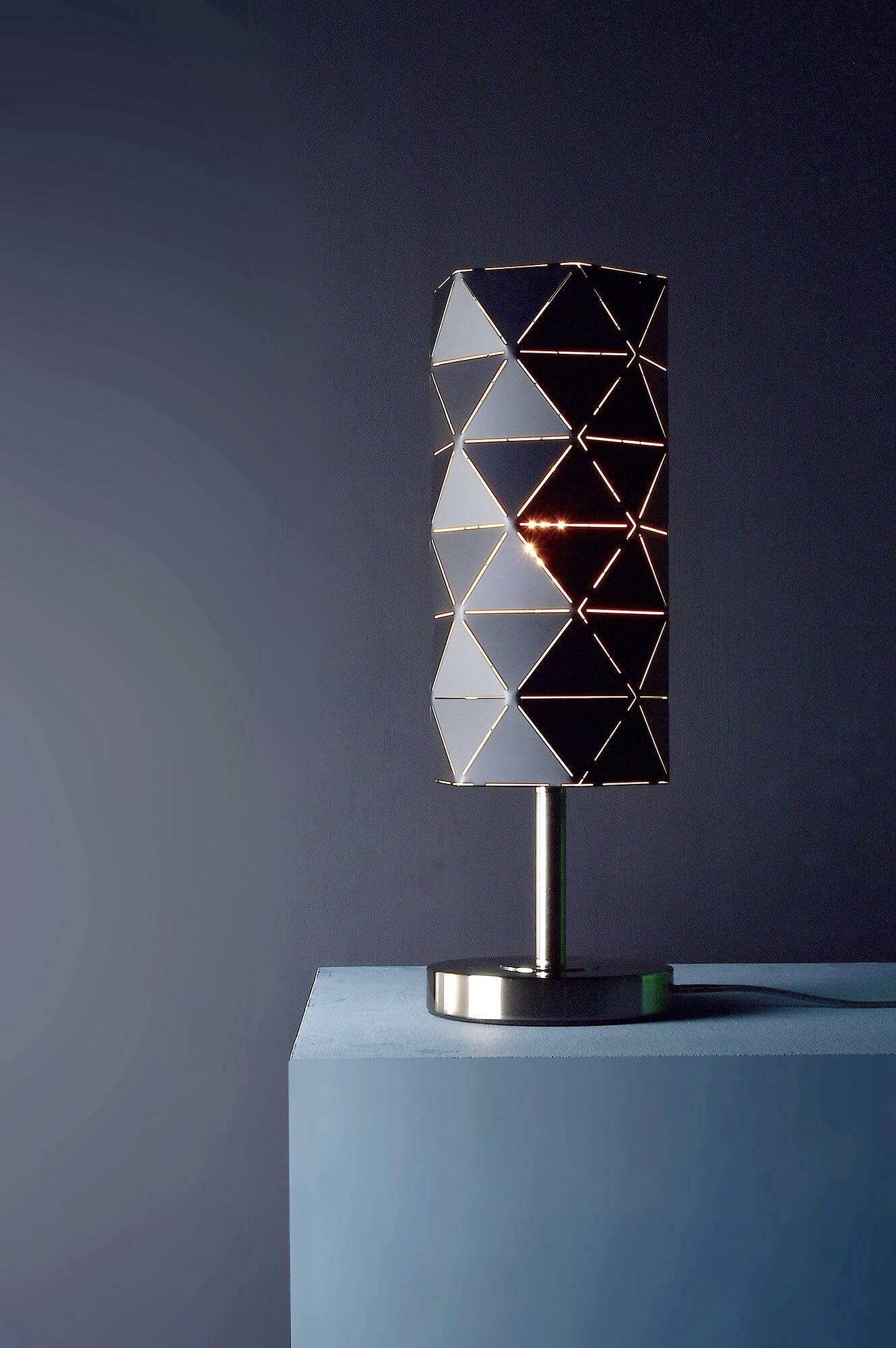 Eine moderne Tischlampe mit geometrischem Muster steht auf einem blauen Tisch vor einem dunkelgrauen Hintergrund.
