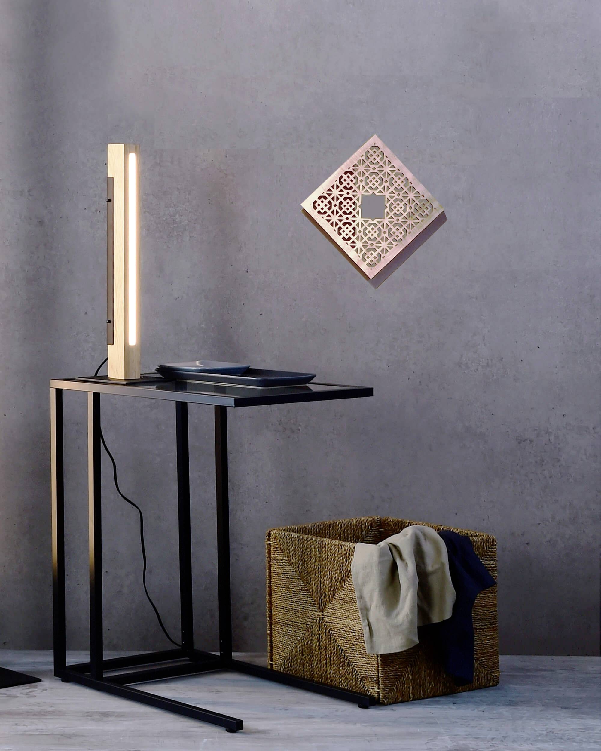Ein minimalistischer Schreibtisch mit einer Lampe und einem Tablett daneben. An der Wand hängt ein dekorativer Spiegel. Ein Korb mit Kleidung steht darunter.