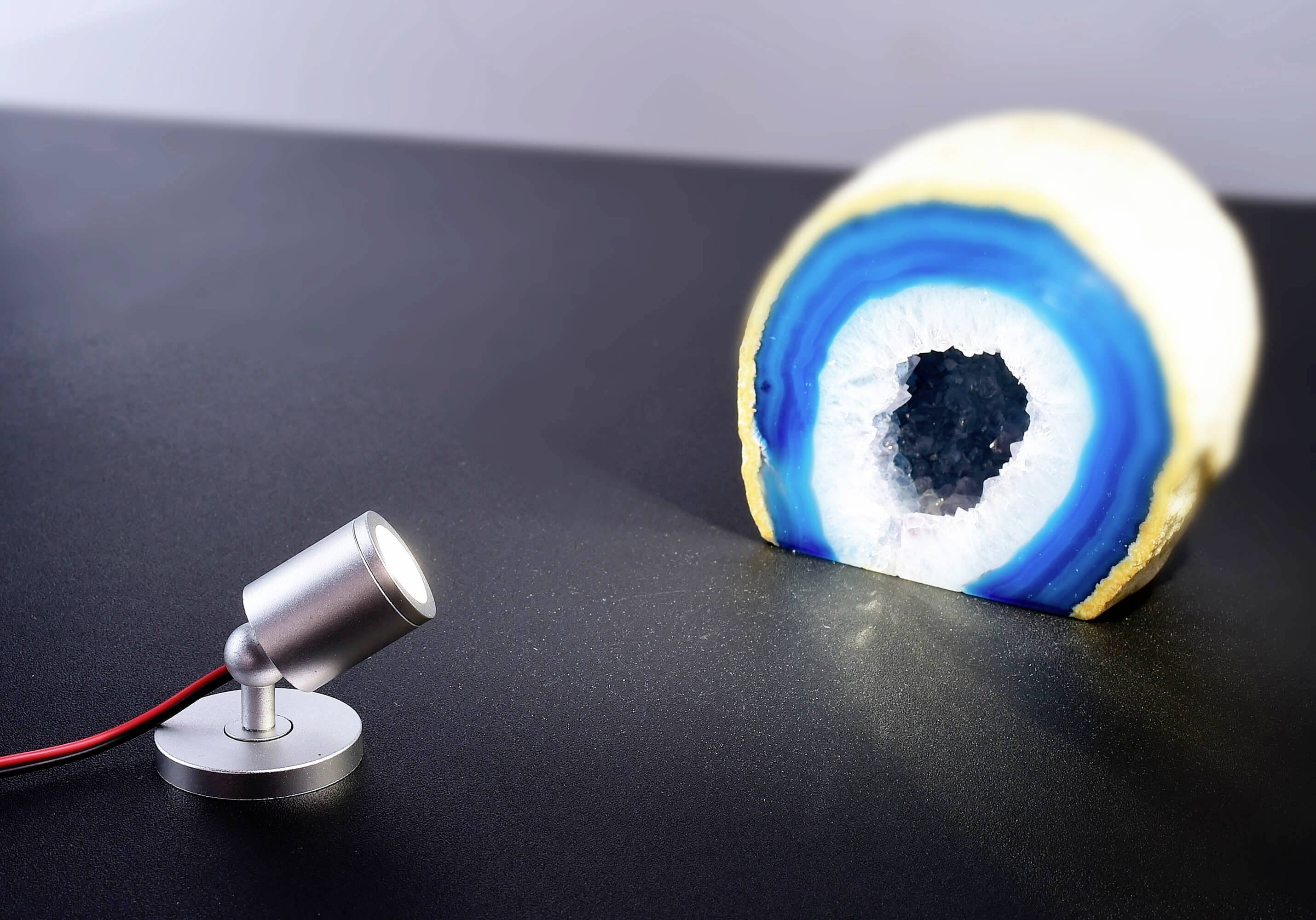 Eine halbe Geode mit blauen und weißen Schichten wird von einem kleinen Strahler beleuchtet, der Schatten auf die Oberfläche wirft.