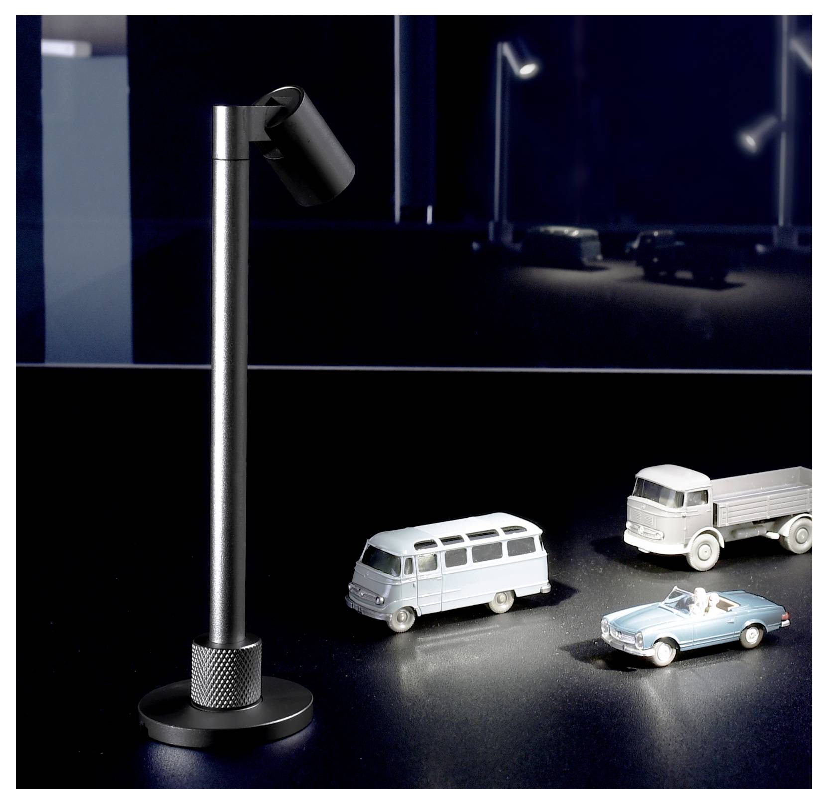 Miniatur-Straßenlaterne neben drei Spielzeugfahrzeugen auf einer dunklen, spiegelnden Oberfläche, mit einem verschwommenen, gespiegelten Hintergrund.