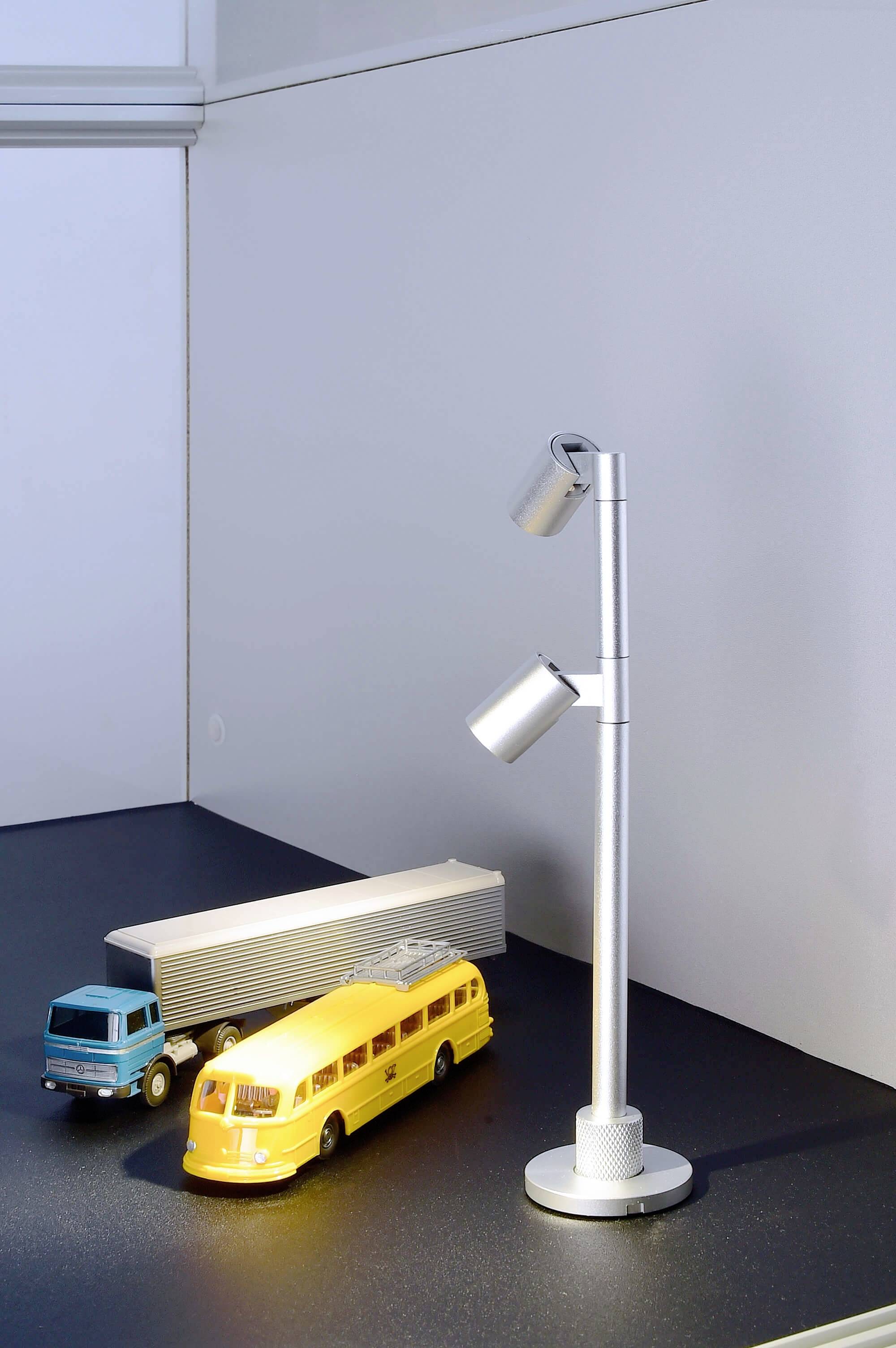 Ein silberner Strahler beleuchtet ein Miniaturmodell mit gelbem Bus, blauem Lastwagen und einem Anhänger in einer Ausstellung.