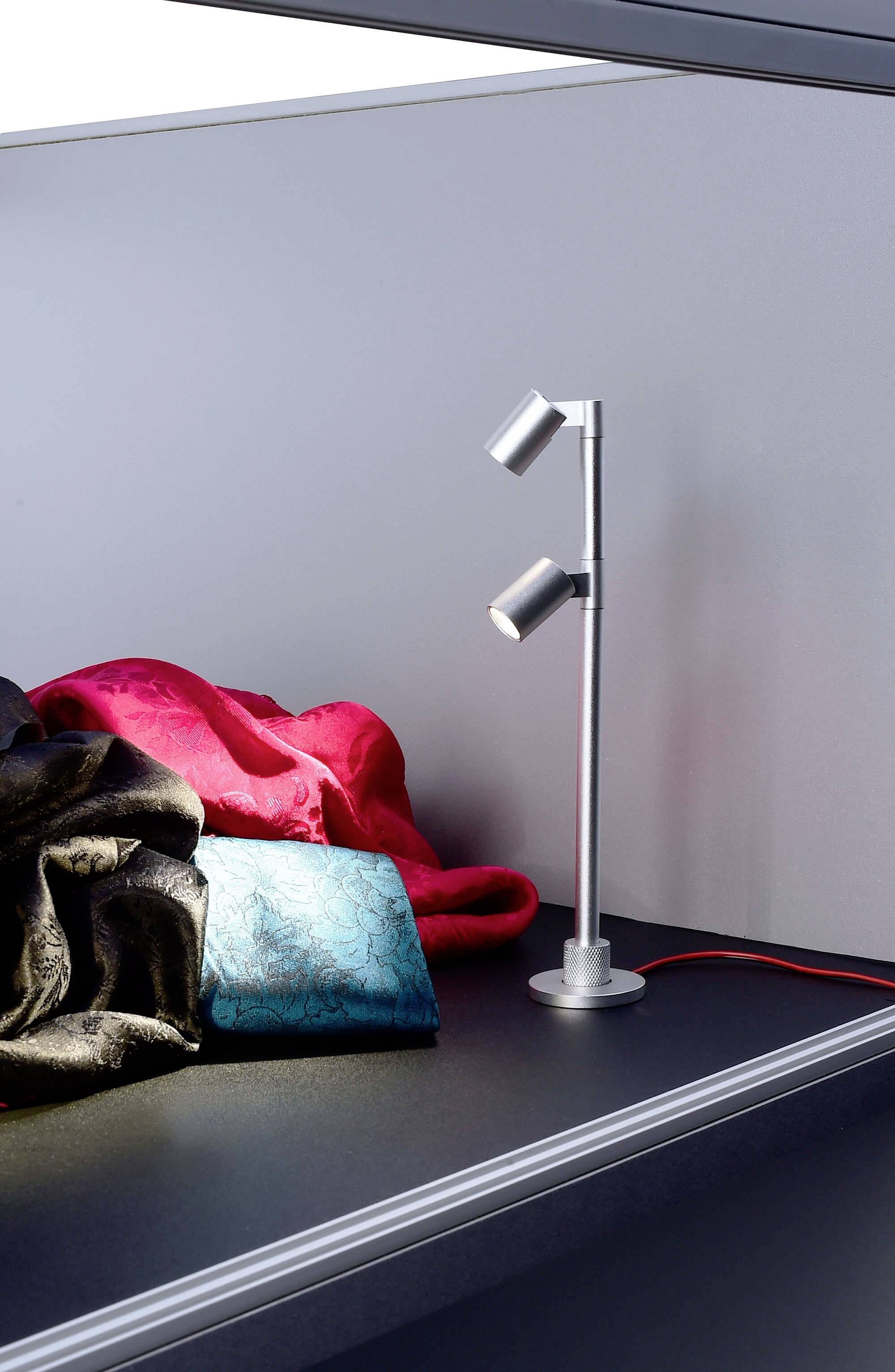 Eine moderne Tischlampe mit zwei Leuchten steht auf einem Tisch. Daneben liegt ein zusammengelegter roter und ein blauer Stoff.