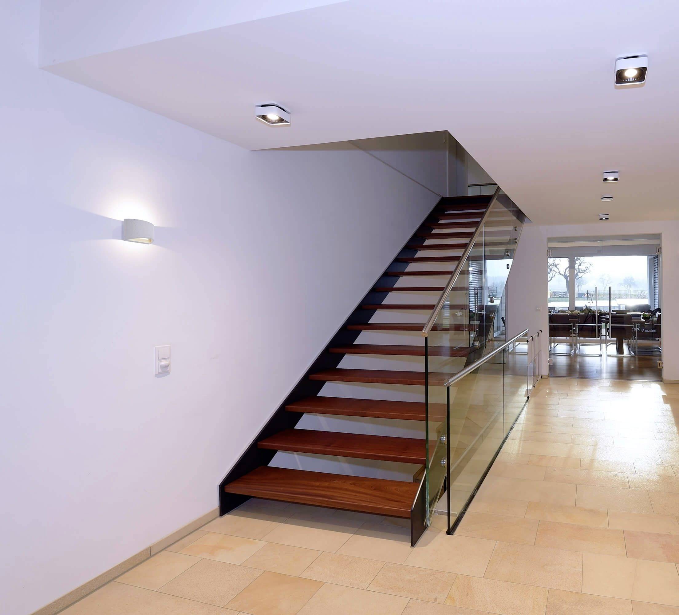Eine moderne Treppe mit Holzstufen und Glasgeländer führt zu einem oberen Bereich. Im Hintergrund ist ein Essbereich zu sehen.