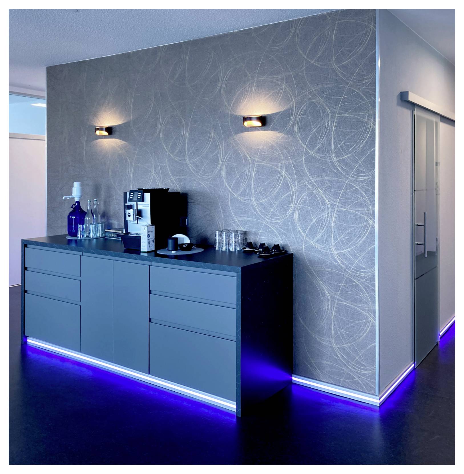 Moderne Küchenzeile mit blauer Unterbauleuchtung, Schränken, Kaffeemaschine, Flaschen und Gläsern vor einer gemusterten grauen Wand in einem Flur.