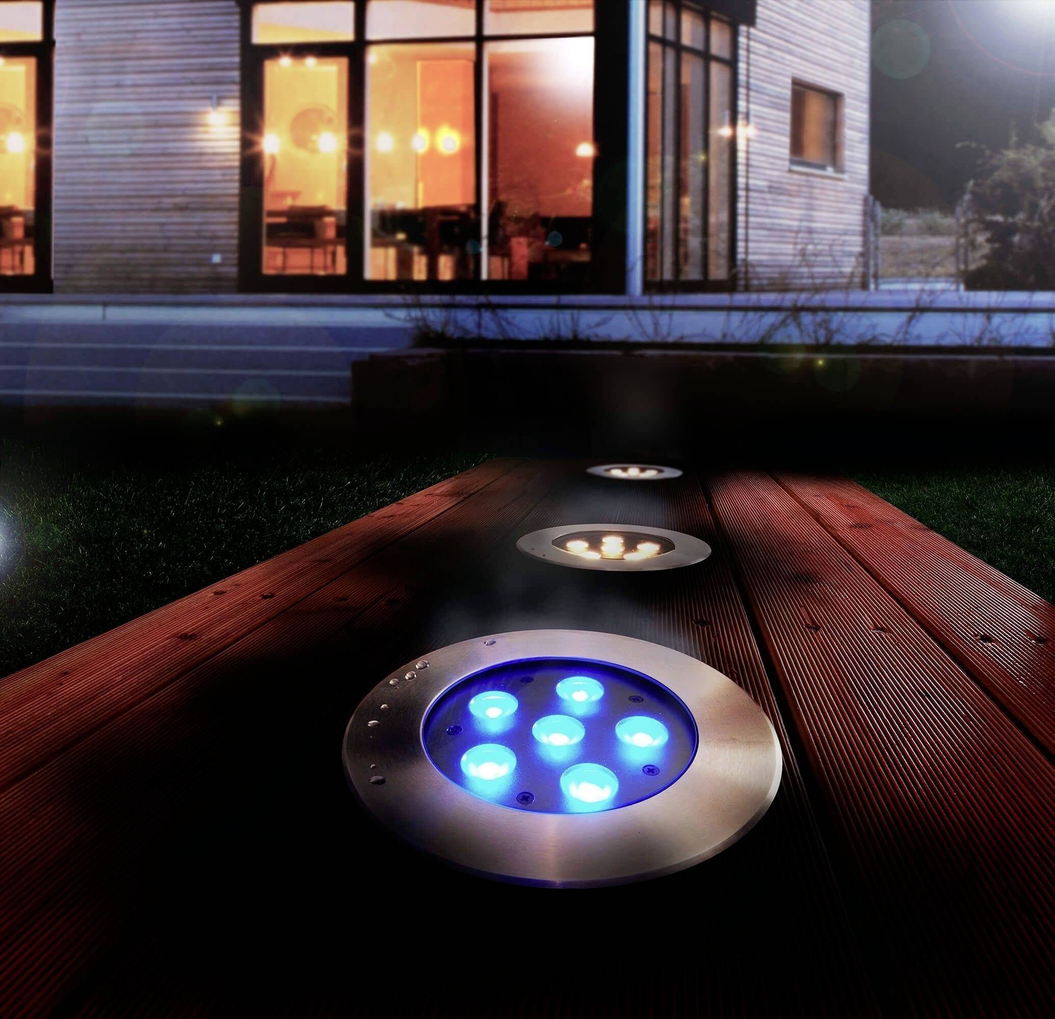 Bodenleuchten strahlen blaues Licht auf einer Holzterrasse aus. Im Hintergrund ist ein modernes, beleuchtetes Haus zu sehen.