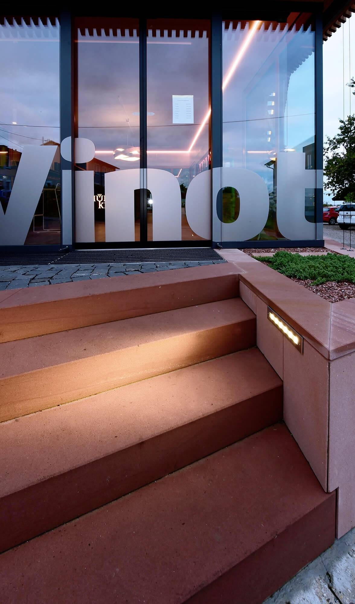 Ein moderner Eingangsbereich mit breiter Treppe führt zu einem Gebäude mit großer Glasfront. Auf der Tür steht 'Vino'.