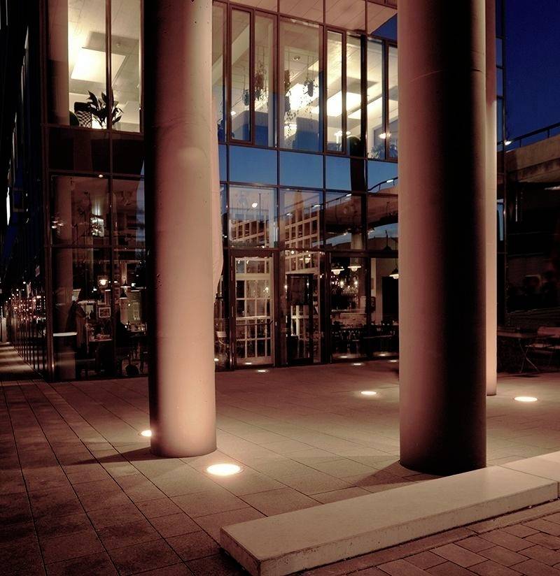 Modernes Gebäude mit großer Glasfassade, beleuchtet bei Dämmerung. Innen sind Lichter und Möbel sichtbar, außen stehen massive Säulen.