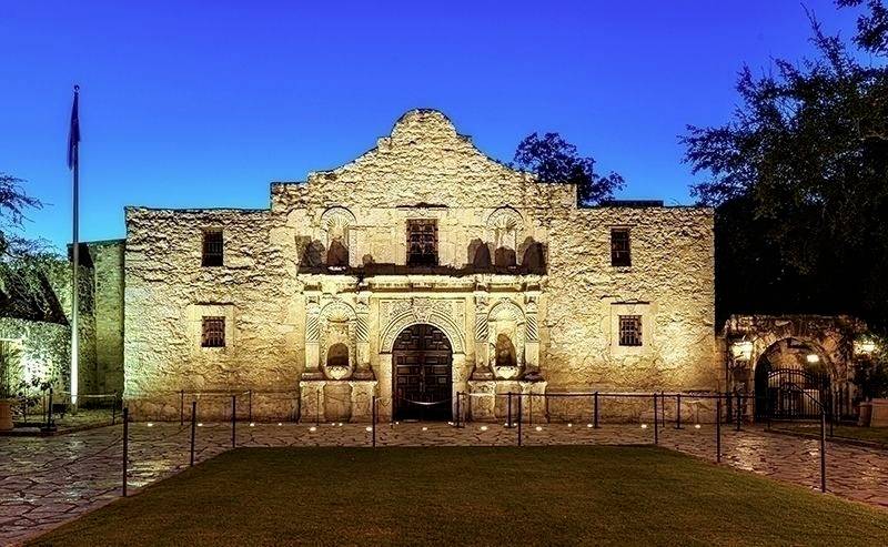 Historisches Gebäude aus Stein mit Rundbögen und Zinnen, beleuchtet bei Nacht. Im Vordergrund ein Rasenstück, im Hintergrund Dämmerung.
