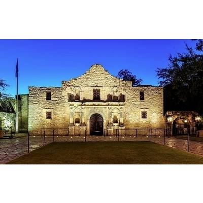 Historisches Gebäude aus Stein mit Rundbögen und Zinnen, beleuchtet bei Nacht. Im Vordergrund ein Rasenstück, im Hintergrund Dämmerung.