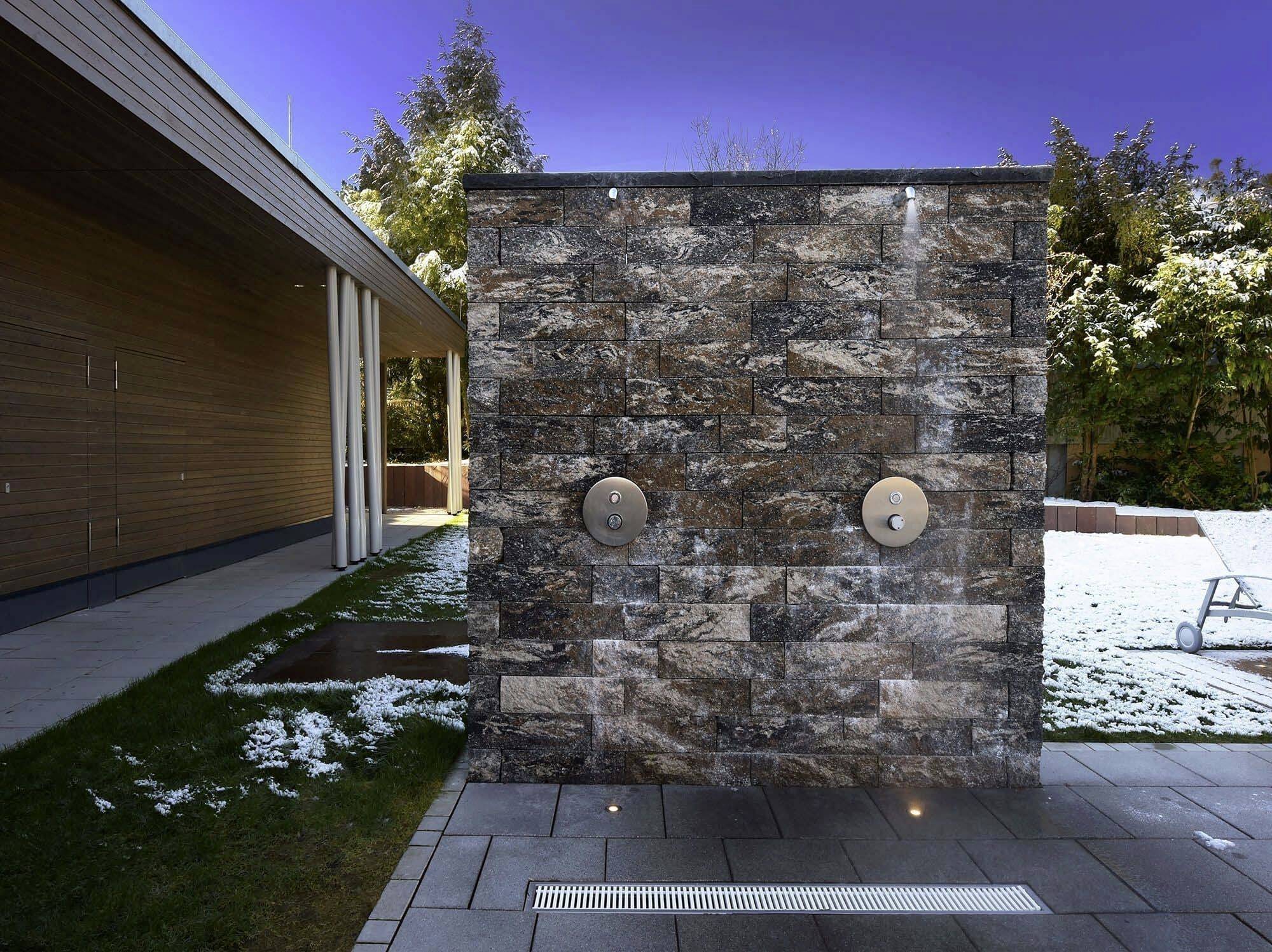 Steinmauer im Garten mit zwei runden Duschen, umgeben von Schnee und einem modernen Gebäude links; klare, kalte Atmosphäre.