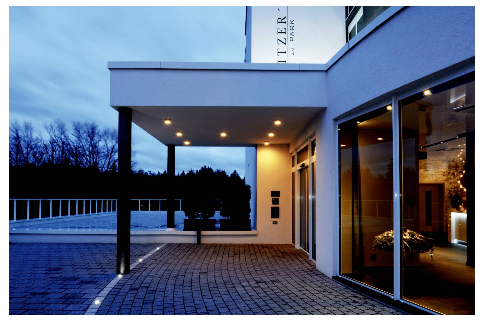 Moderner Gebäudeeingang in der Dämmerung mit beleuchtetem überdachtem Gehweg, Glastüren und gepflasterem Innenhof.