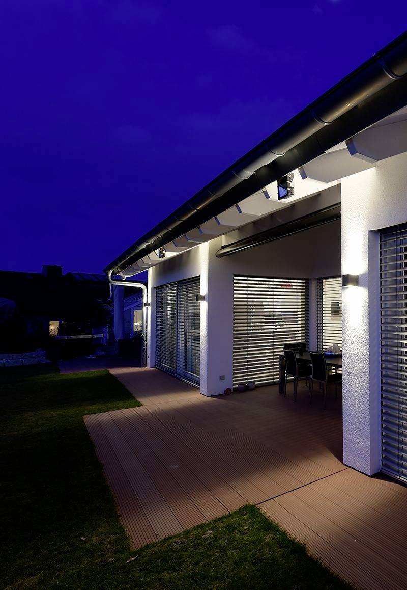 Ein modernes Haus mit beleuchtetem Terrassenbereich und großen Fenstern in der Abenddämmerung. Tisch und Stühle im Freien sichtbar.