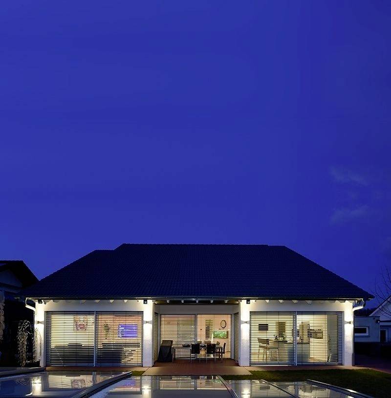 Ein modernes Einfamilienhaus bei Nacht, beleuchtet mit großen Fenstern, durch die ein Wohnzimmer und Essbereich erkennbar sind.