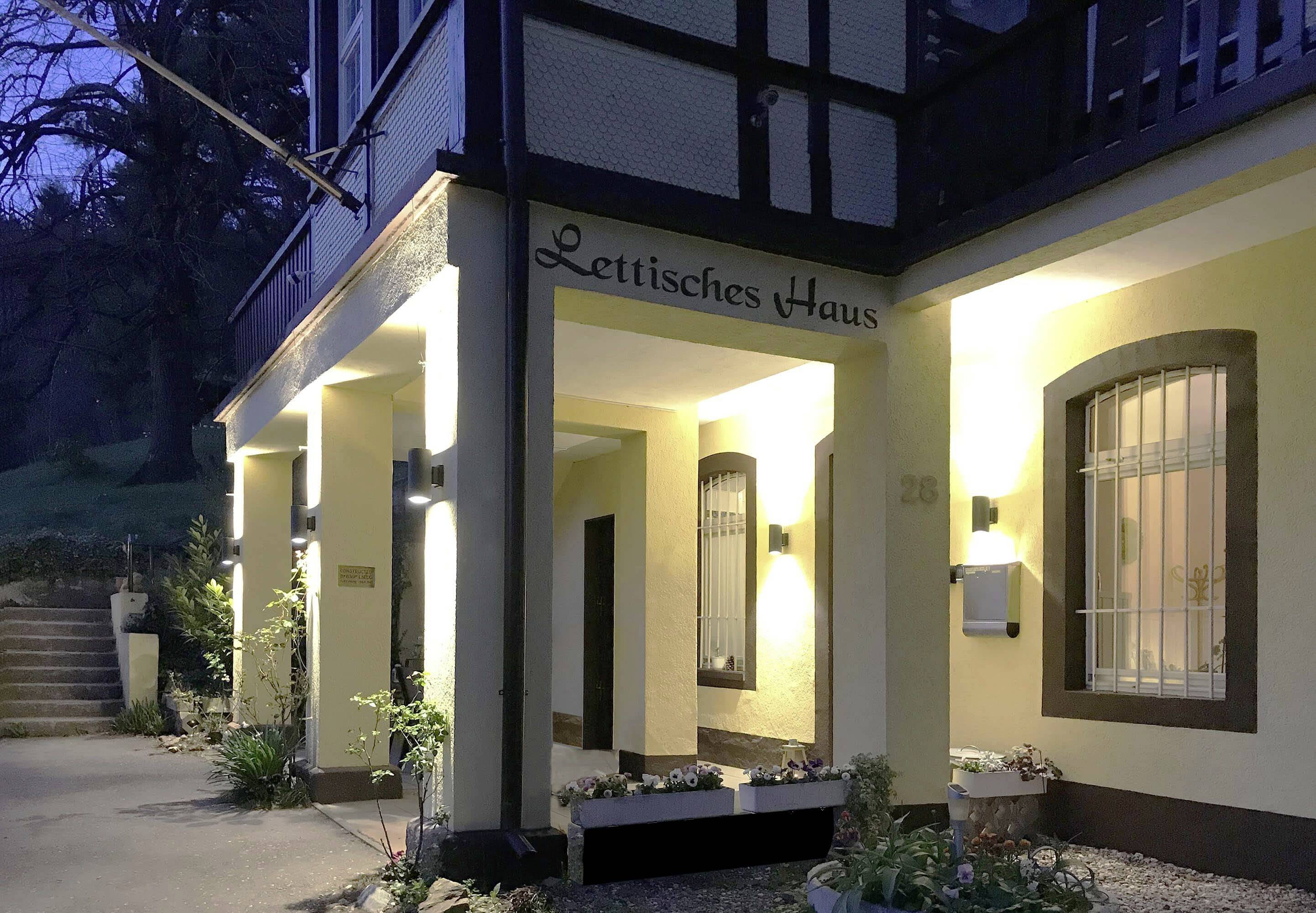 Eingang eines Hauses bei Dämmerung, beleuchtet. Beschriftung über dem Eingang: 'Lettisches Haus'. Pflanzen dekorieren den Bereich.
