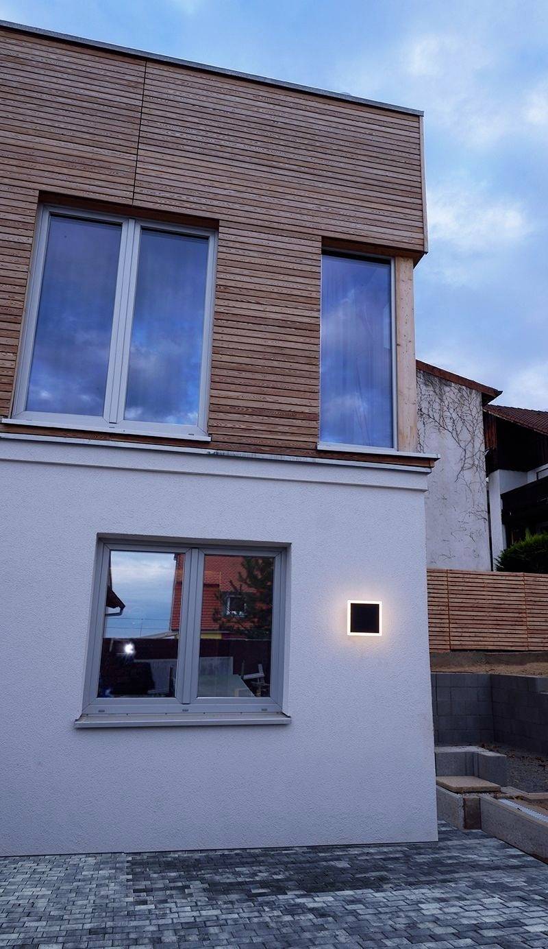 Zweistöckiges modernes Haus mit Holzverkleidung und großen Fenstern, beleuchtet im Abendlicht. Pflastersteine führen zum Eingang.