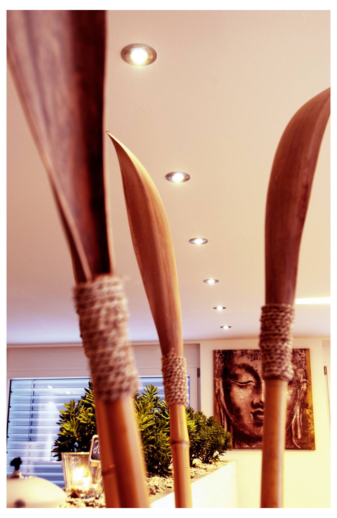 Spa- oder Wellnessraum mit hohen, schilfartigen Holzskulpturen im Vordergrund und einem Buddha-Porträt an der Wand im Hintergrund.