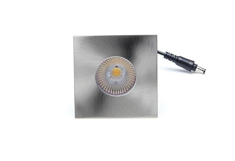 Quadratische LED-Leuchte mit Metallfinish und sichtbarem Leuchtmittel in der Mitte, schwarze Anschlussbuchse rechts.