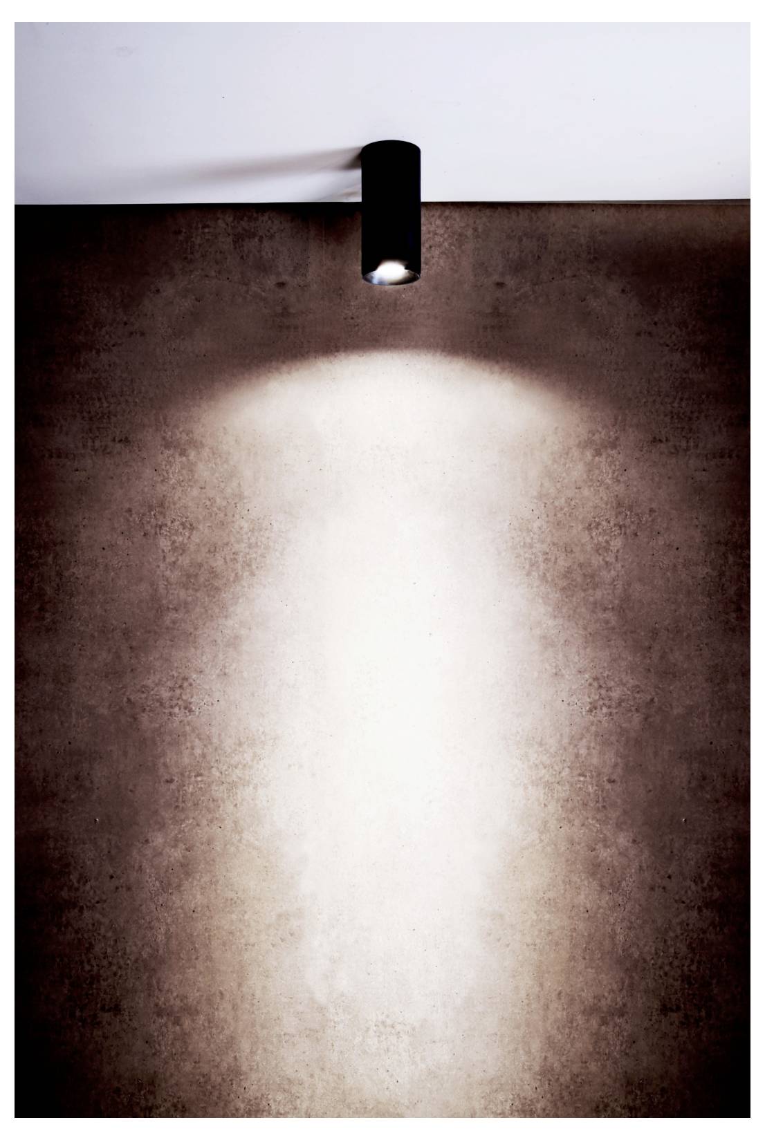 Ein wandmontierter Strahler wirft einen hellen Lichtkegel an einer braun strukturierten Wand hinab und erzeugt so eine dramatisch beleuchtete Mitte.