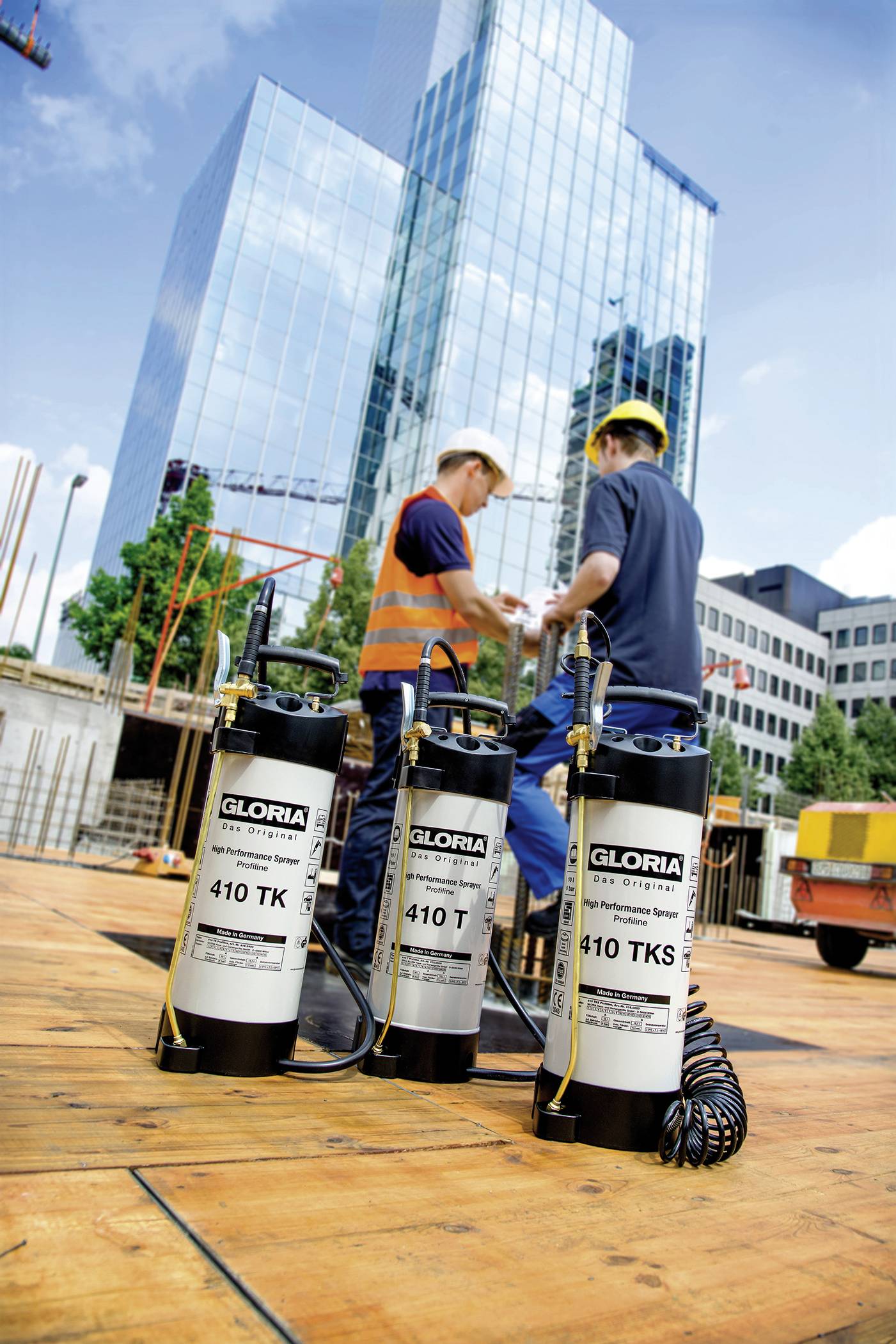 Zwei Bauarbeiter auf einer Baustelle besprechen Pläne; im Vordergrund stehen drei Feuerlöscher; im Hintergrund moderne Bürogebäude.