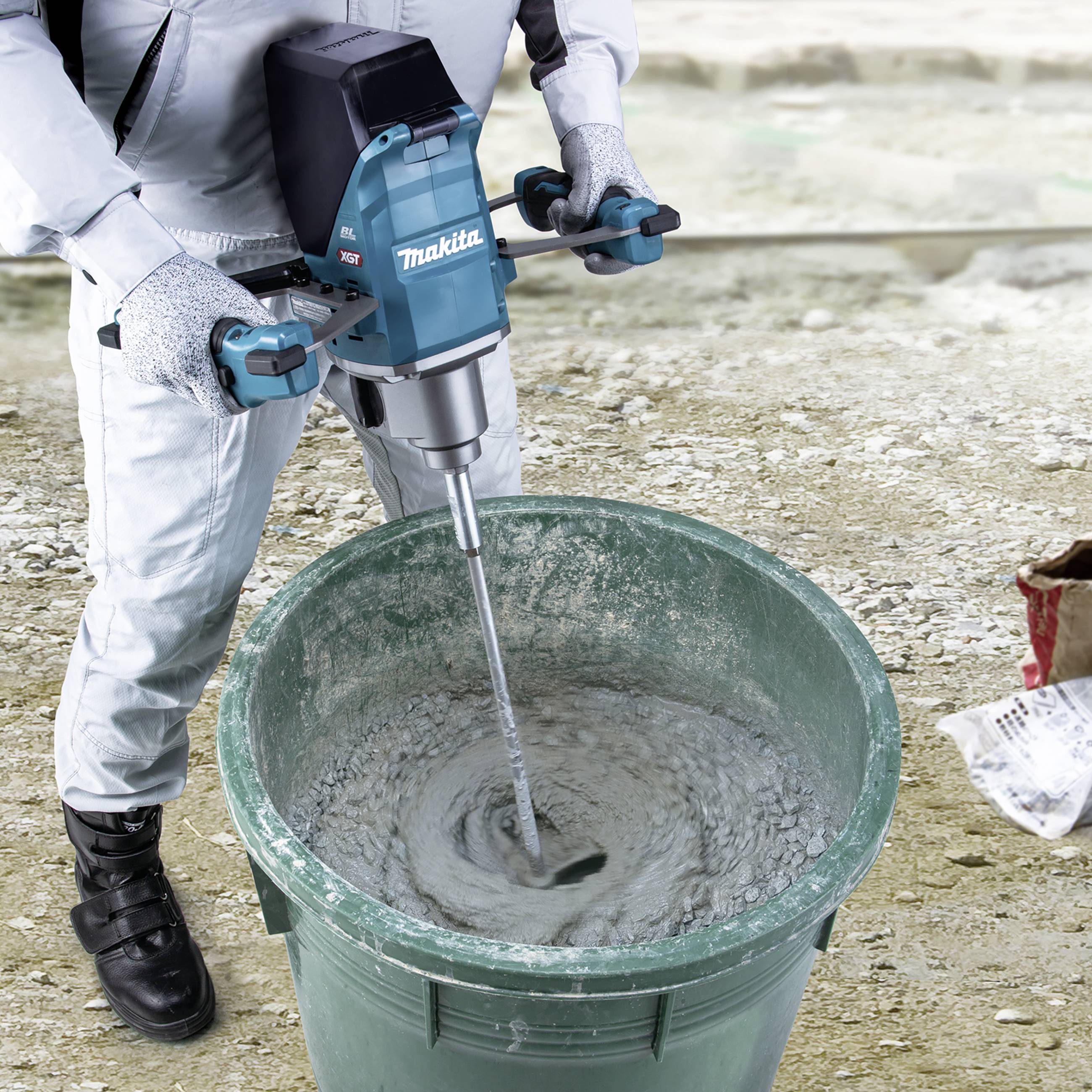 Eine Person mischt Beton in einem grünen Eimer mit einem elektrischen Rührgerät von Makita. Die Person trägt Schutzkleidung.