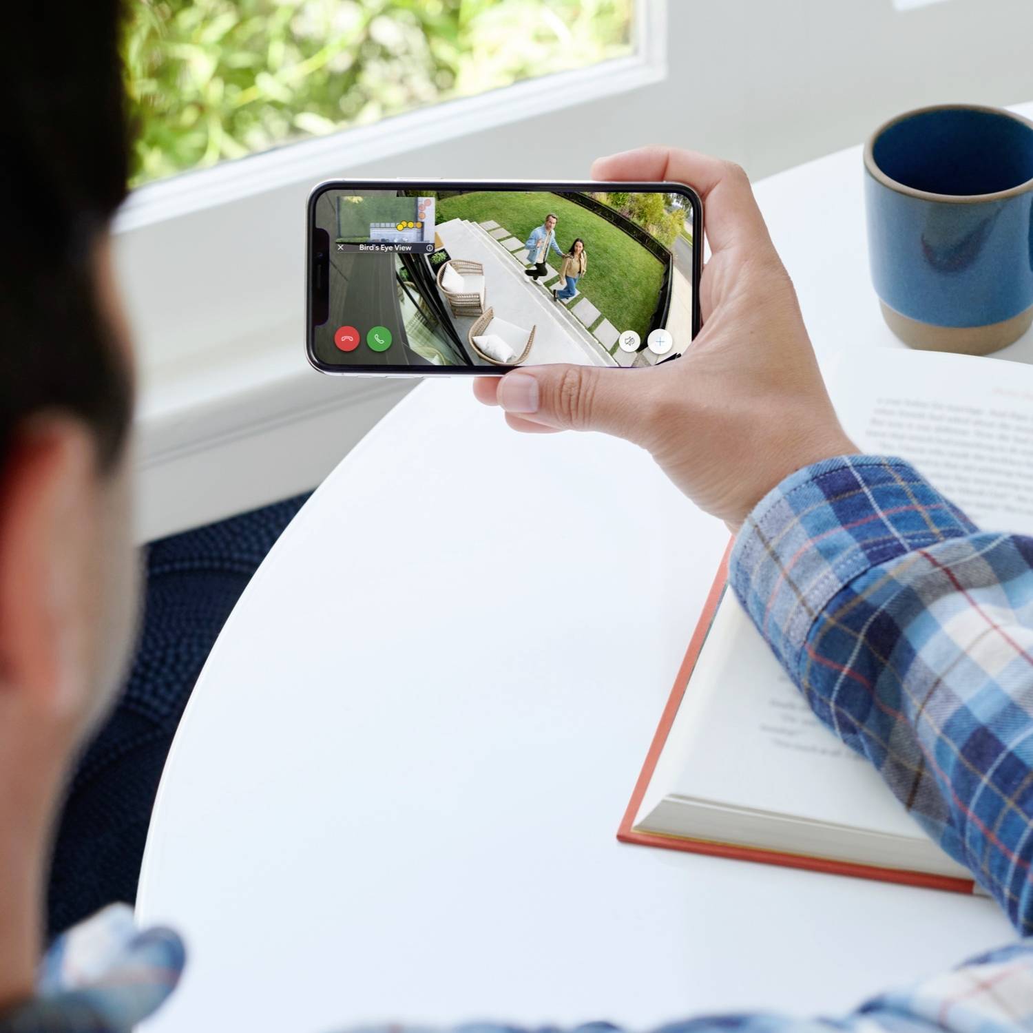 Eine Person hält ein Smartphone, das ein Überwachungsbild eines Gartens zeigt. Auf einem Tisch daneben liegt ein geöffnetes Buch.