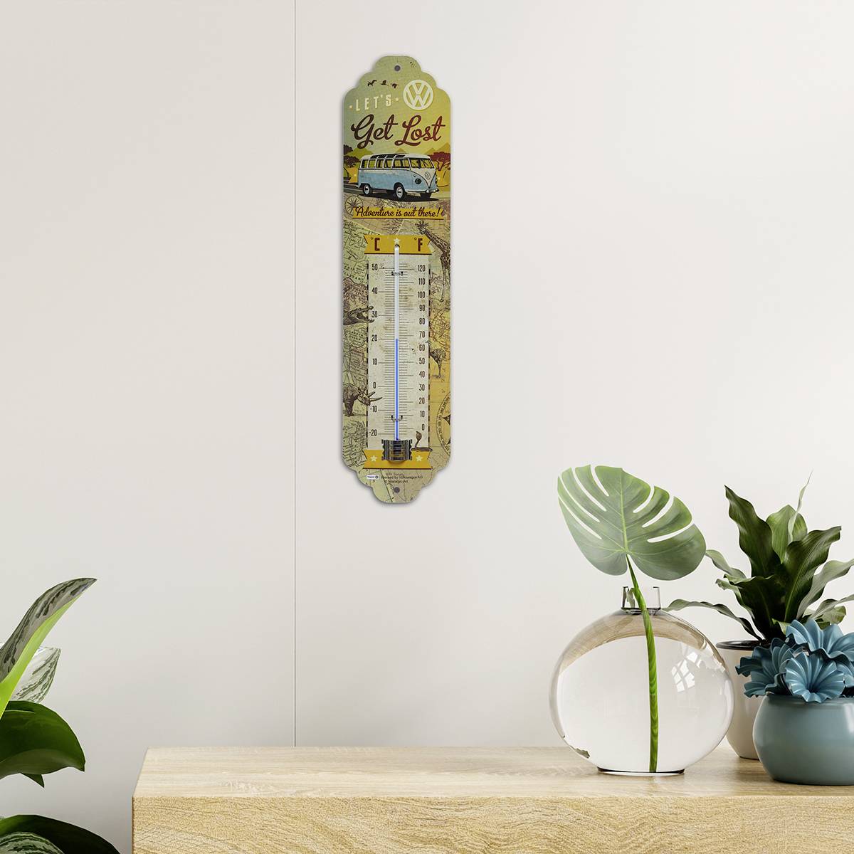 Wandthermometer mit VW-Design und 'Let's Get Lost'-Aufschrift über einem Regal. Pflanzen links und rechts ergänzen die Dekoration.