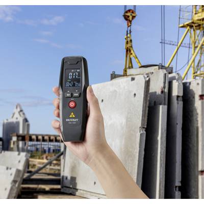 Eine Hand hält ein digitales Messgerät vor einer Baustelle mit Betonplatten und Kränen im Hintergrund.