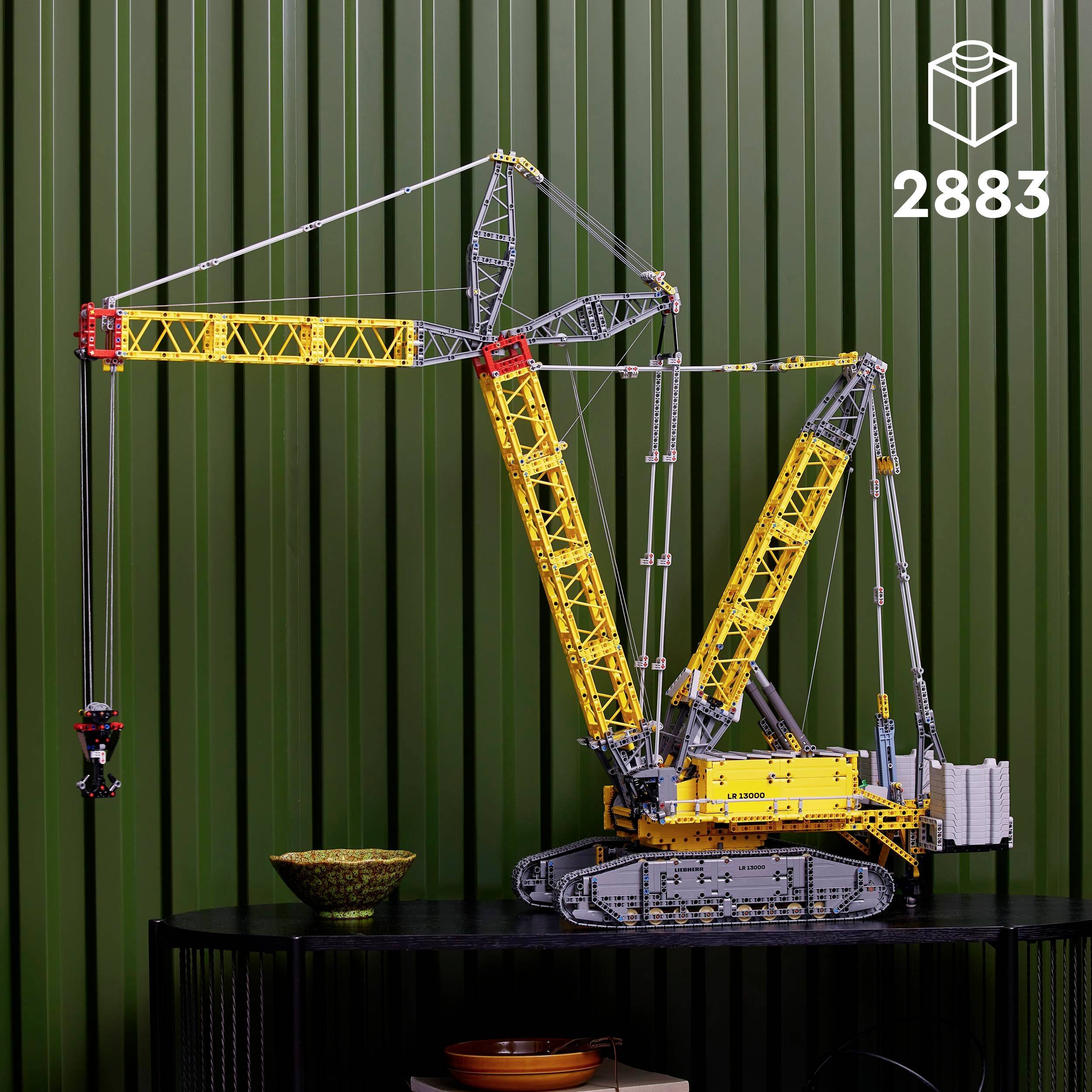 Spielzeugmodell eines gelben Baukrans aus Legosteinen, aufgebaut auf einem Tisch vor einer grünen Wand. Set-Nummer '2883'.