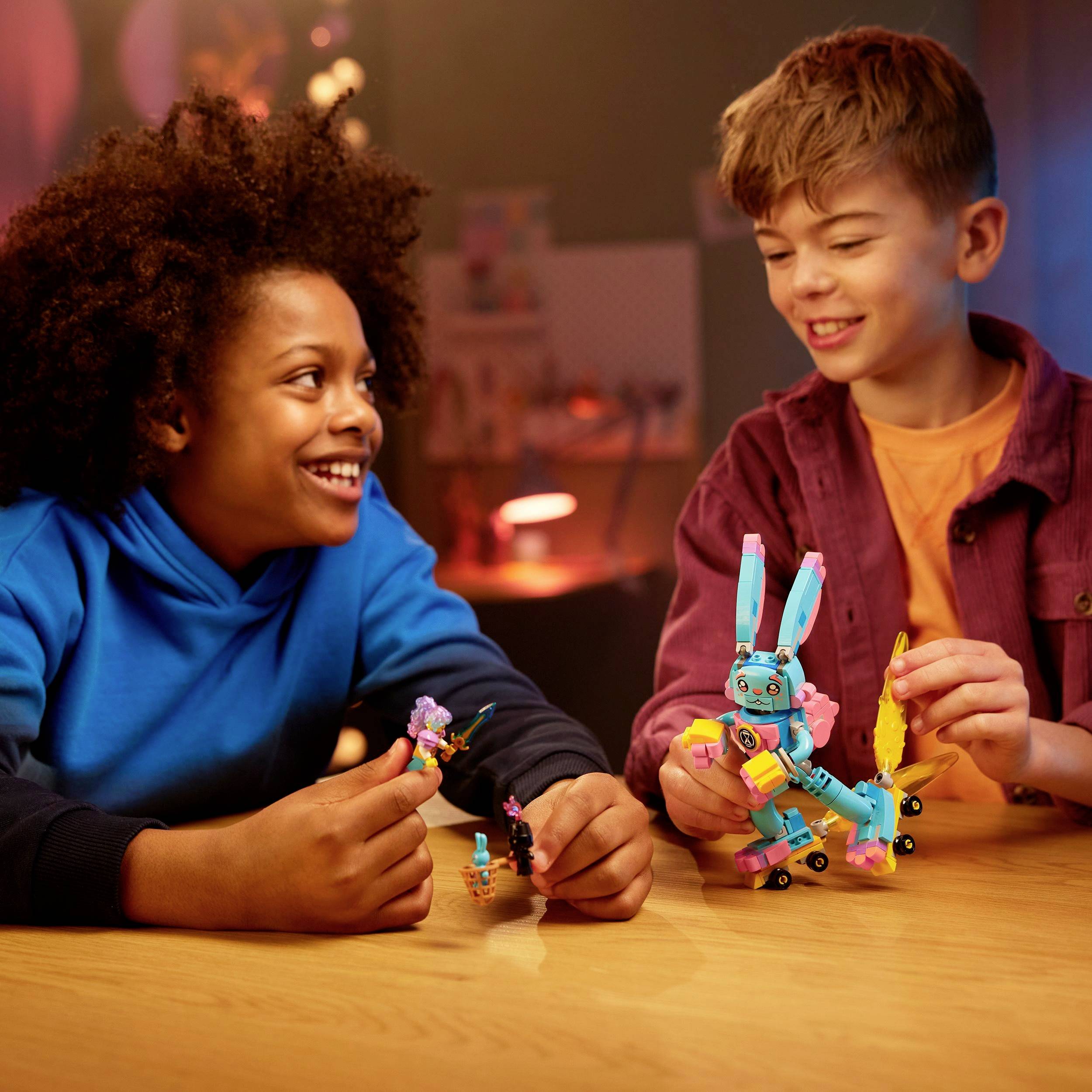 Zwei Kinder sitzen an einem Tisch und spielen mit einem bunten Roboter-Spielzeug. Sie lächeln und scheinen Freude am Spiel zu haben.