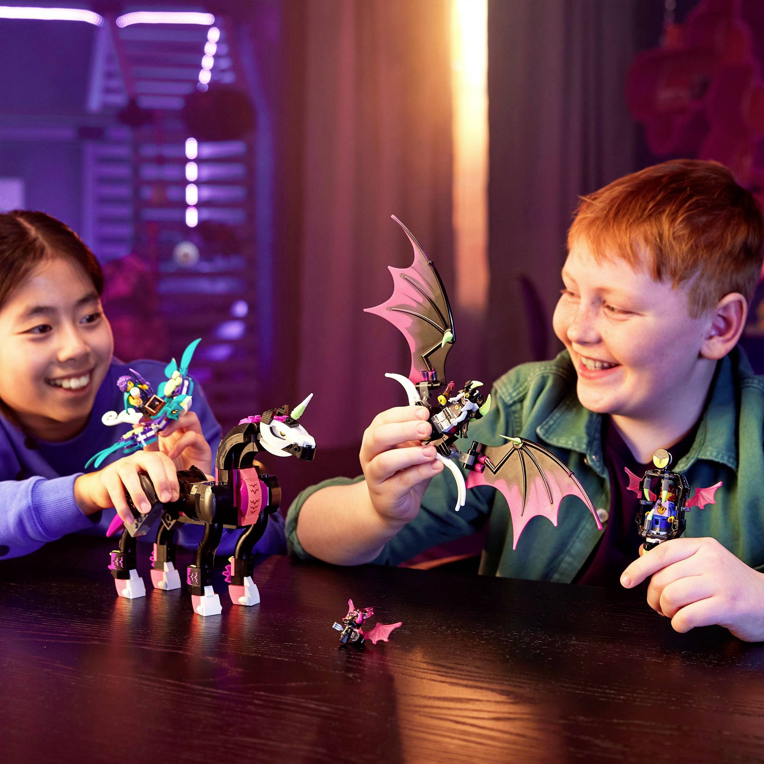 Zwei Kinder sitzen an einem Tisch und spielen lachend mit bunten Spielzeugfiguren und einem Pferd. Die Szene ist bunt beleuchtet.