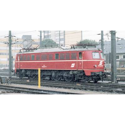 Rote Elektrolokomotive steht auf Bahngleisen. Hintergrund zeigt industrielle Gebäude und Stromleitungen.