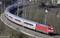 Ein roter Zug fährt auf einer kurvigen Bahnstrecke durch eine ländliche Landschaft mit Bäumen und vereinzelten Häusern im Hintergrund.
