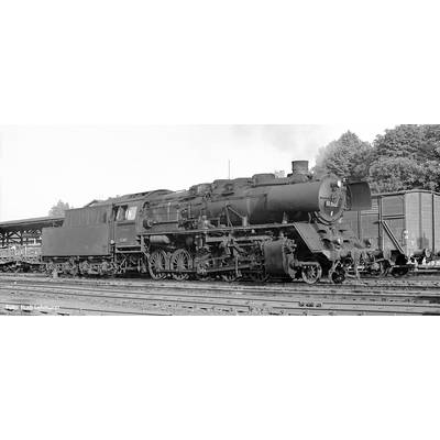 Eine Dampflokomotive steht auf einem Bahngleis. Im Hintergrund sind ein Bahnhof und Bäume sichtbar. Die Lokomotive ist groß und massiv.