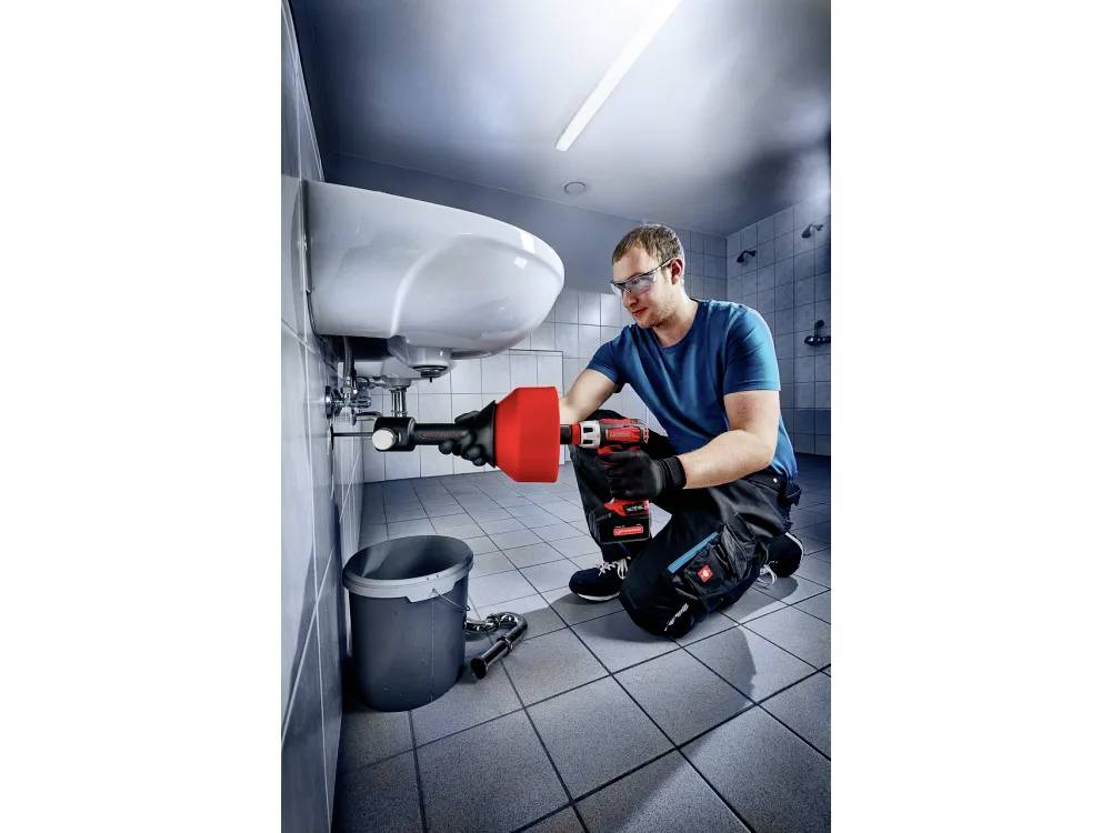 Ein Klempner mit Schutzausrüstung verwendet ein Werkzeug, um ein Rohr unter einem Waschbecken zu reparieren. Eine Werkzeugbox und ein Eimer befinden sich auf dem gefliesten Boden in der Nähe.