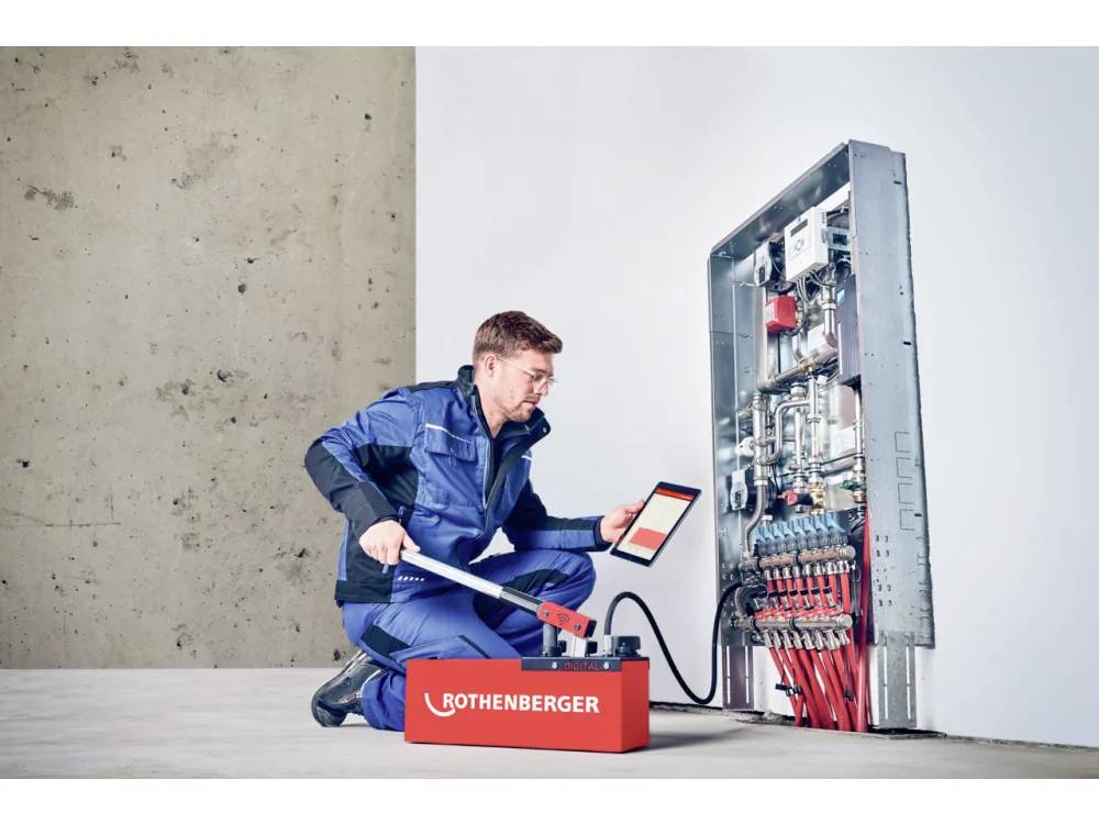 Ein Techniker in blauen Overalls verwendet ein hydraulisches Werkzeug mit einem Digitaltablett, um ein an der Wand montiertes Panel mit Rohren und Ventilen zu inspizieren.