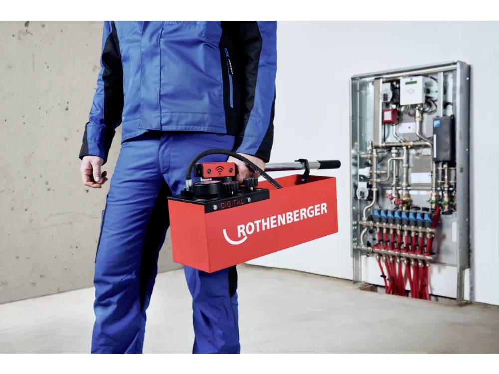 Eine Person in blauer Arbeitskleidung trägt eine rote Rothenberger-Maschine in einem Raum mit freigelegter Sanitärausrüstung, was auf eine Wartungsumgebung hindeutet.