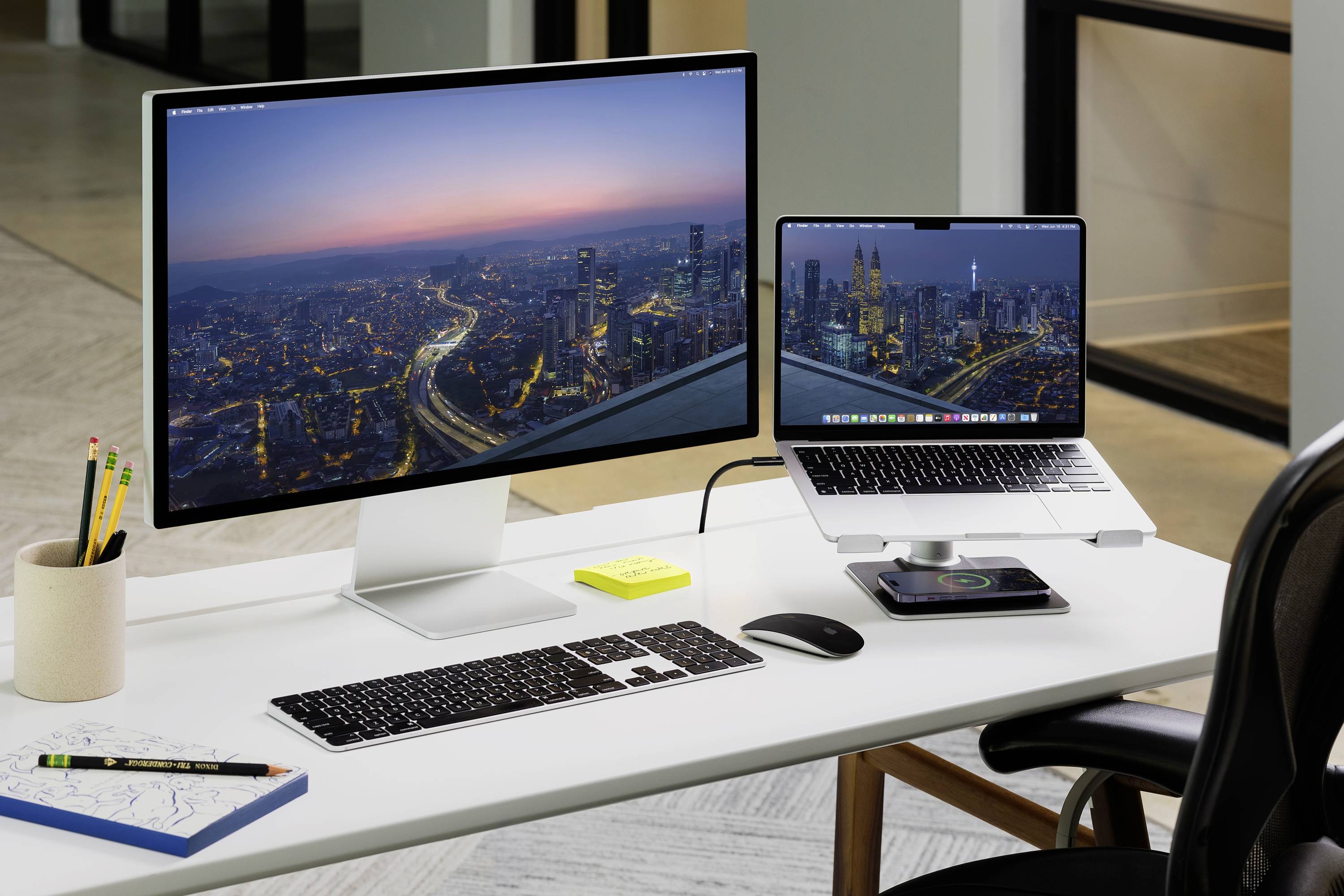 Ein Schreibtisch mit einem großen Monitor links und einem Laptop rechts auf einem Ständer. Beide Bildschirme zeigen eine Stadtansicht bei Dämmerung.