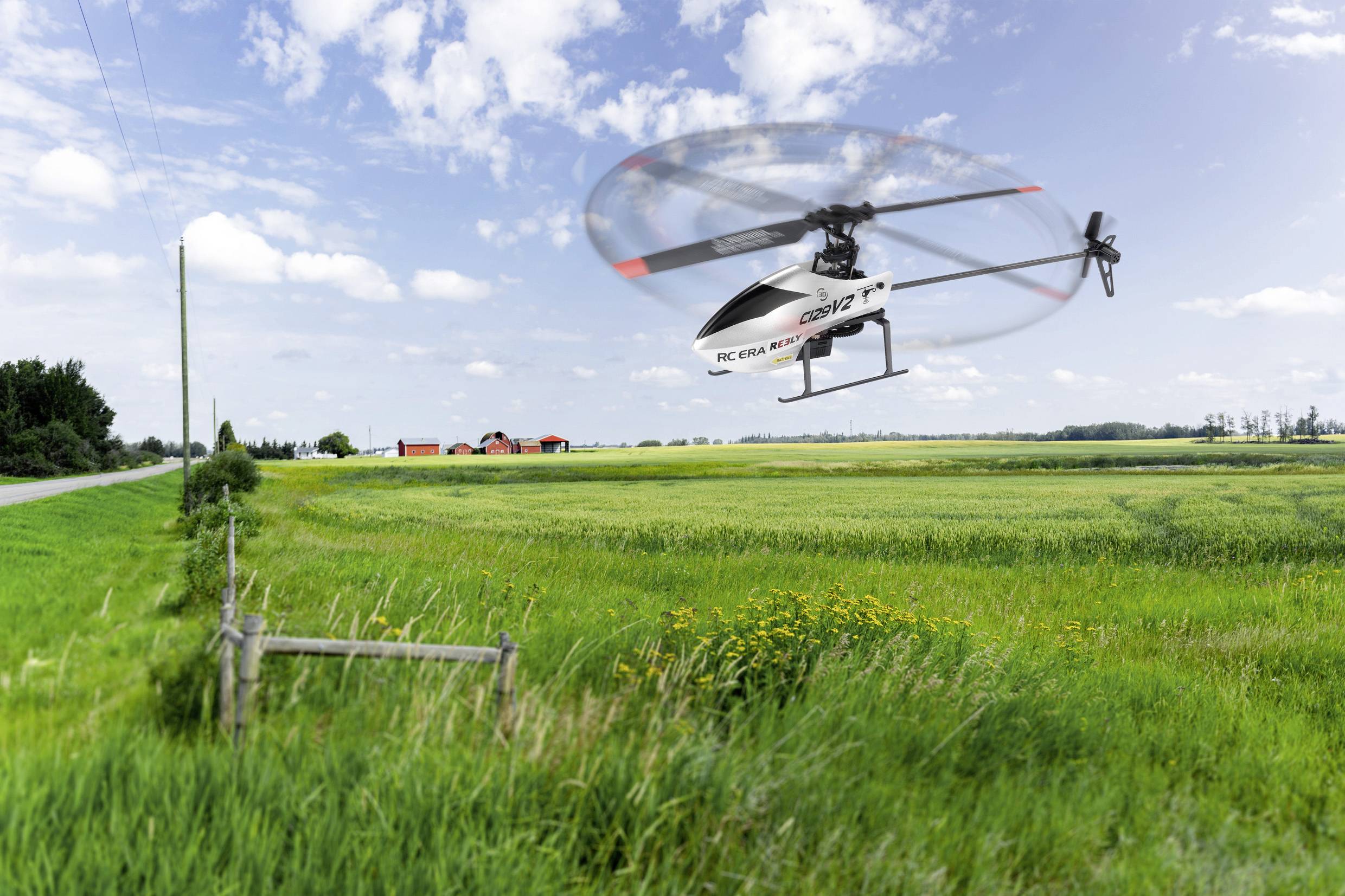 Ein ferngesteuerter Hubschrauber fliegt über ein grünes Feld mit einem rot-weißen Bauernhof im Hintergrund unter einem bewölkten Himmel.