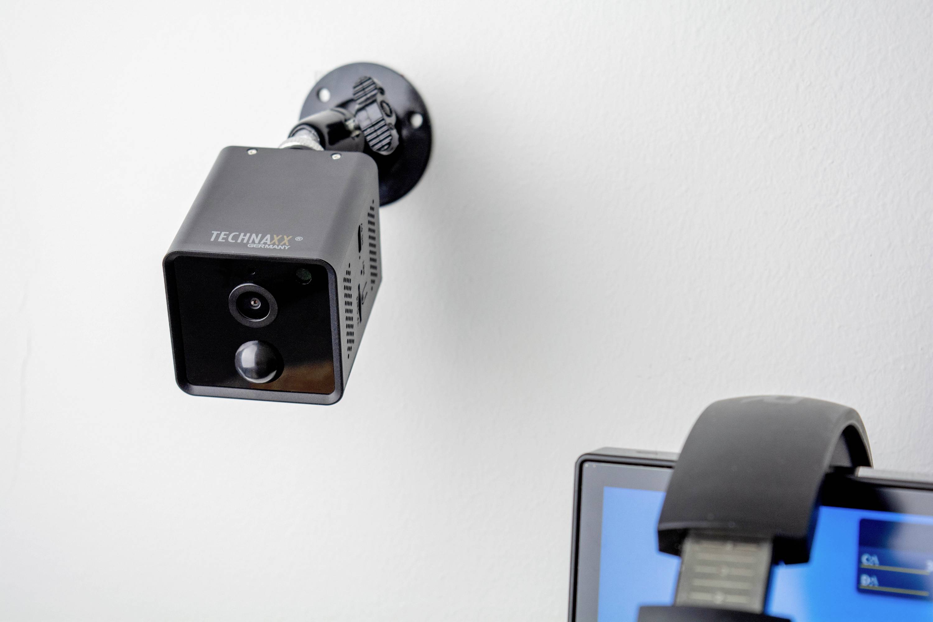 Überwachungskamera an der Wand neben einem Bildschirm mit Kopfhörern. Die Kamera ist schwarz und quadratisch und zeigt nach unten.