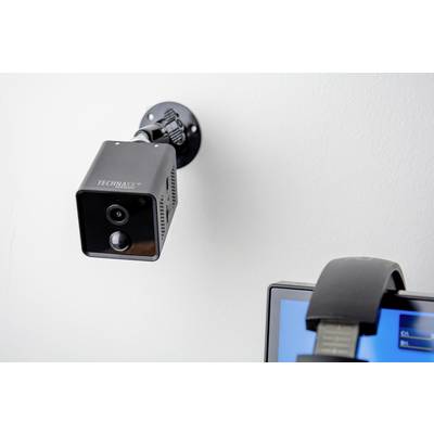 Überwachungskamera an der Wand neben einem Bildschirm mit Kopfhörern. Die Kamera ist schwarz und quadratisch und zeigt nach unten.