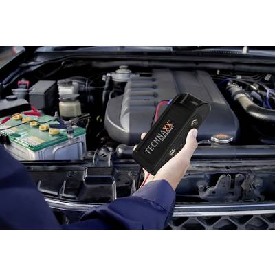 Eine Person hält ein Starthilfegerät nahe einer Autobatterie im Motorraum eines Fahrzeugs.