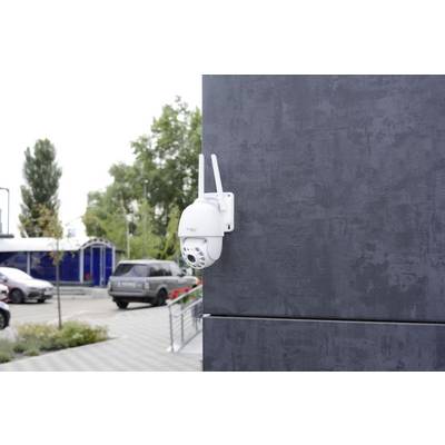 'Überwachungskamera an einer grauen Wand angebracht, mit Blick auf einen Parkplatz mit Autos und Bäumen im Hintergrund.'
