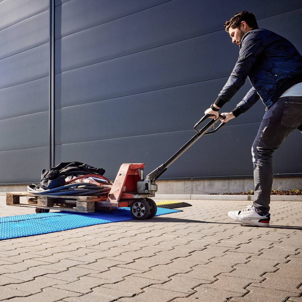 Ein Mann zieht einen Hubwagen mit mehreren Taschen darauf über einen gepflasterten Gehweg vor einem industriellen Gebäudetor.