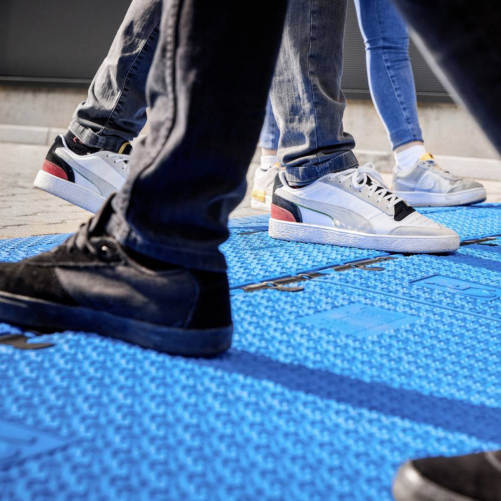Eine Gruppe von Personen geht auf einer blauen, taktilen Bodenfläche. Unterschiedliche Schuhe sind sichtbar, einige mit weißen Sohlen.