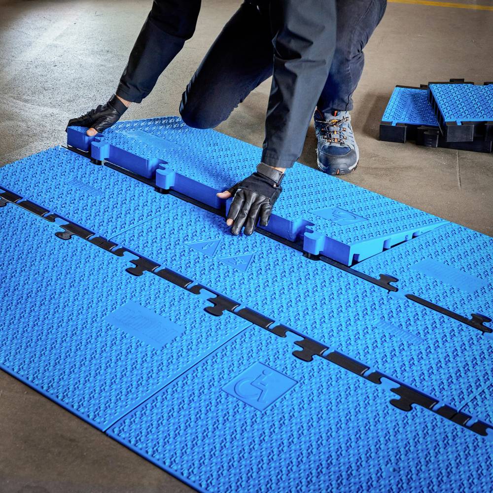 Eine Person montiert blaue Bodenmatten auf einem Werkstattboden. Die Matten haben rutschfeste Oberflächen und passen ineinander.