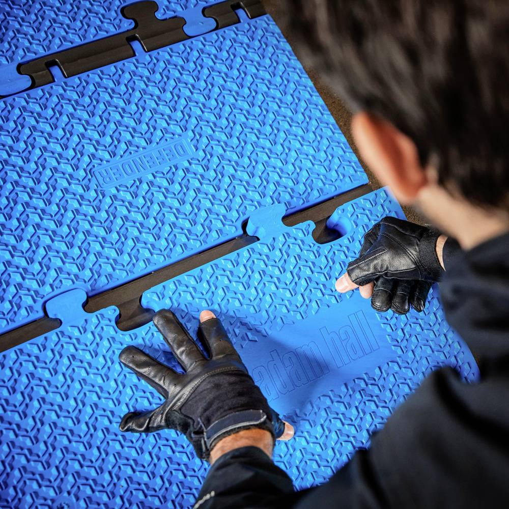 Eine Person mit Handschuhen montiert blaue Bodenplatten mit rutschfester Struktur. Die Platten haben Verzahnungen zum Verbinden.