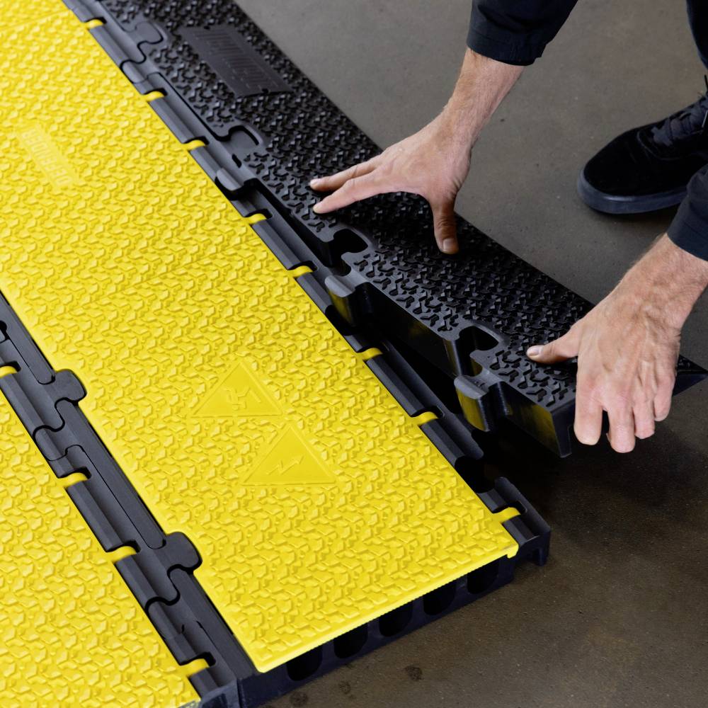 Zwei Hände montieren gelbe Kunststoffbodenplatten mit rutschfester Struktur. Die Platten haben Zapfenverbindungen für einfache Installation.